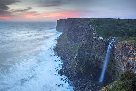 The sun at sunset, illuminates the waves crashing against the cliffs of Capo Nieddu where a magical waterfall plunges into the wonderful sea of Sardinia