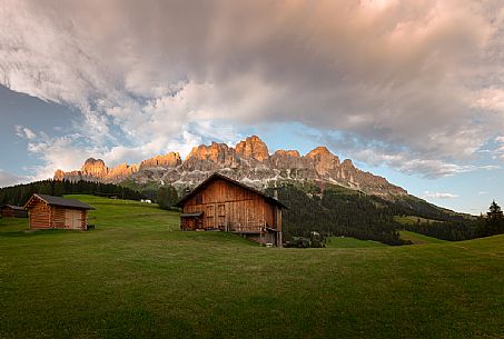 The Catinaccio mountain range at the sunset with some chalet on a green meadow and cloudy orange sky, dolomites, South Tyrol, Italy, Europe